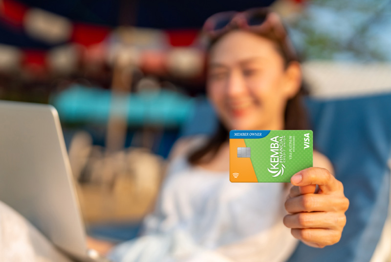 A woman sitting on a chair with a laptop on her lap holding up a member owner KEMBA Visa.