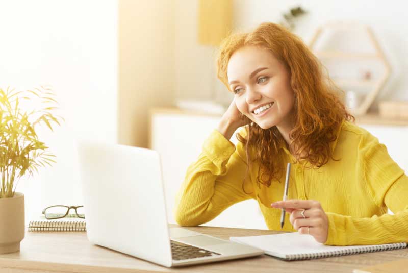 A woman sitting at a laptop and smiling with a pen in her hand.
