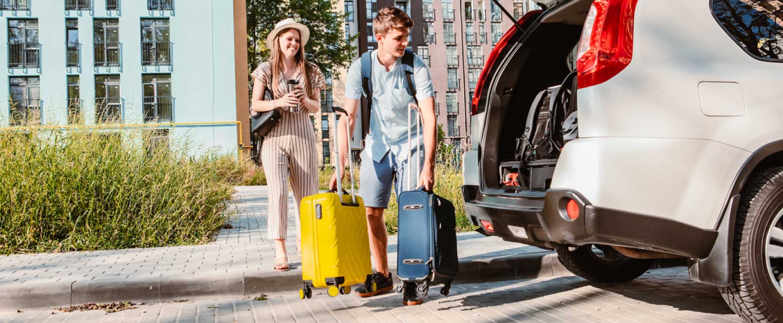 A girl standing on the sidewalk and a guy putting two suitcases into the trunk of a car.
