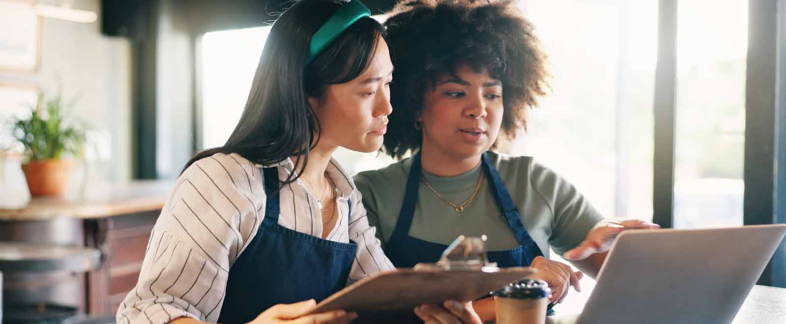 Two café small business owners collaborate using a laptop and clipboard in a bright, modern workspace.