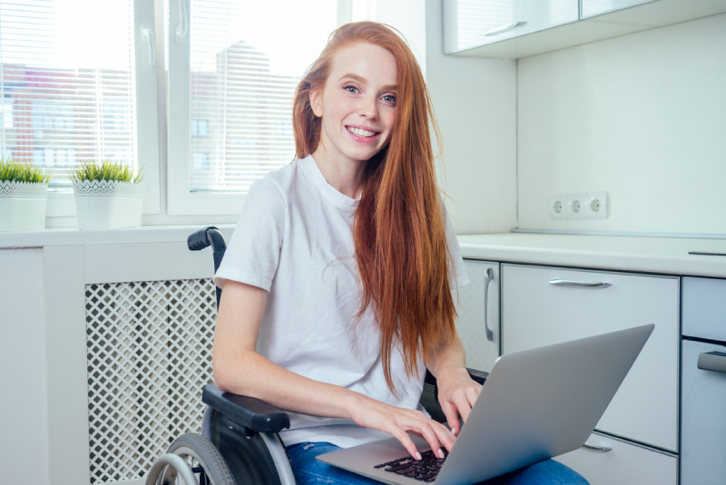 A girl sitting in a wheelchair smiling with a laptop on her lap.