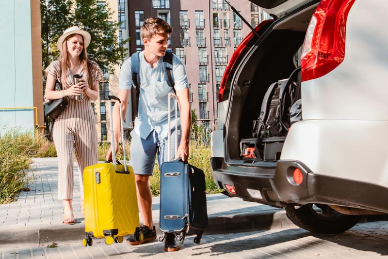 A girl standing on the sidewalk and a guy putting two suitcases into the trunk of a car.
