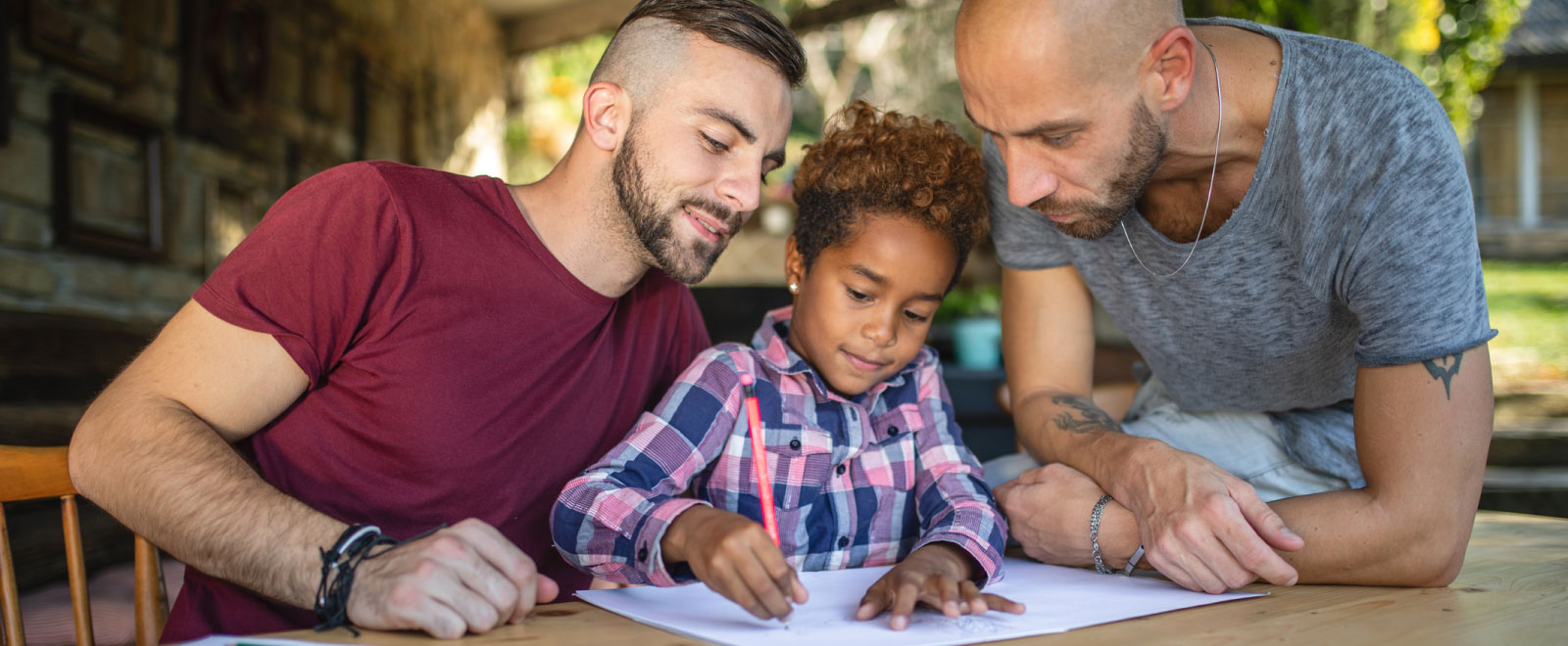 Two men sitting at a table with a child watching her draw.
