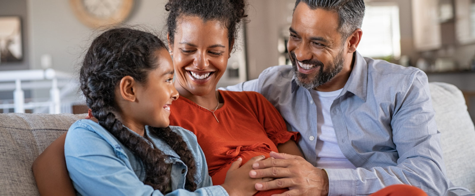 A daughter, mom and dad sitting on a couch their hands on the pregnant mom's stomach.
