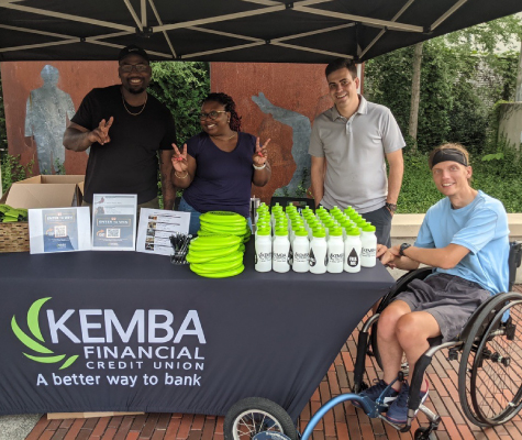 A group of four people at a booth with KEMBA merchandise under a tent.