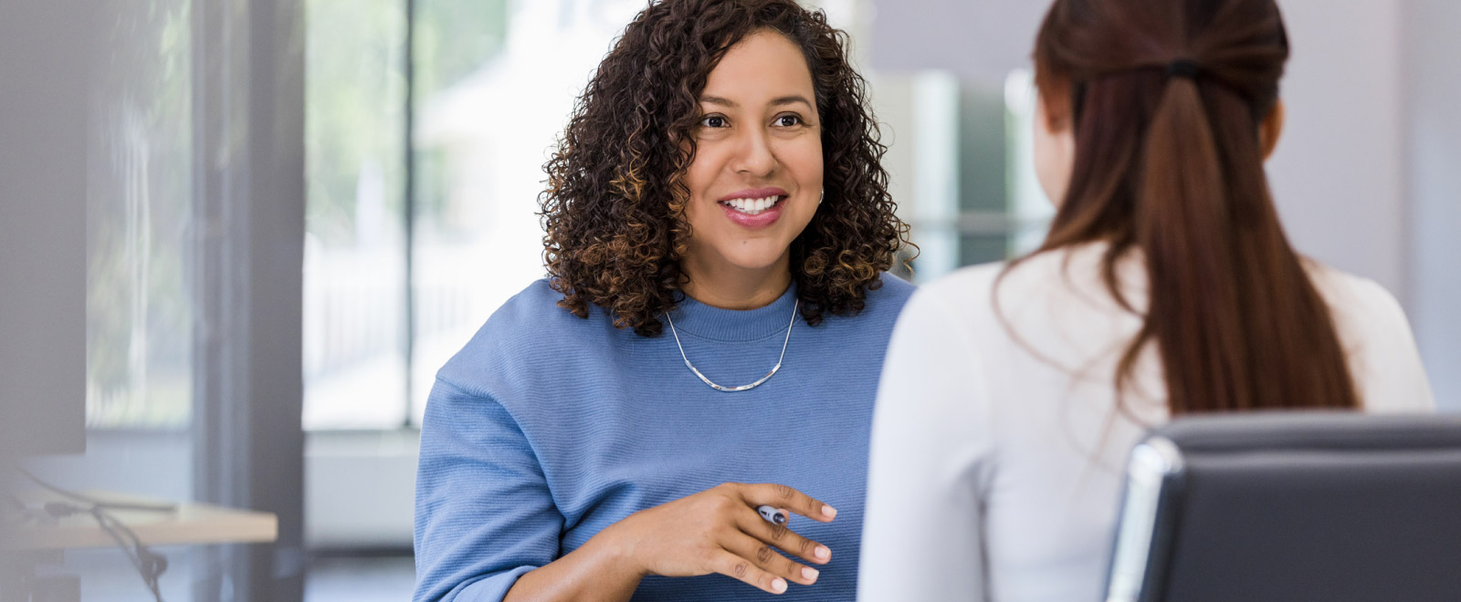 A woman with her back to the camera speaking to a woman across the table that is smiling at her and holding a pen.