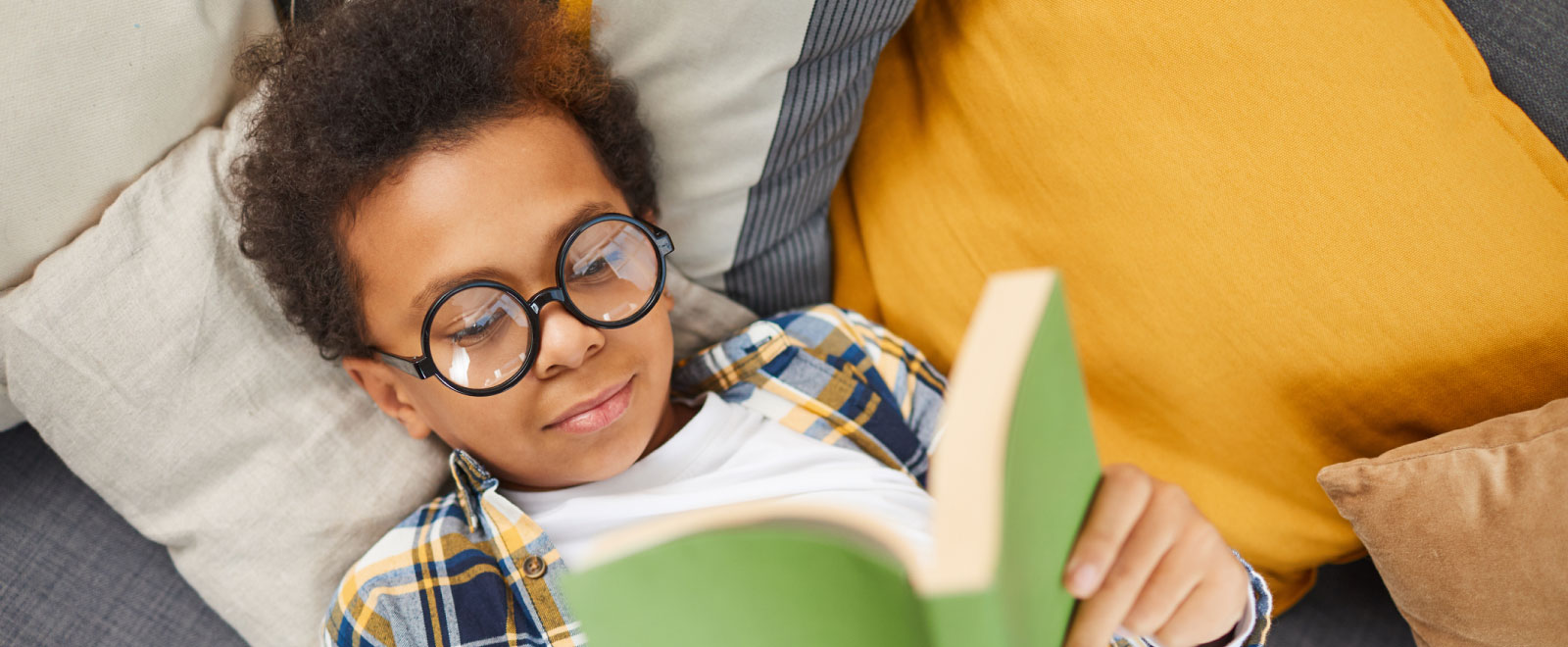 A young boy laying on a couch reading with glasses on.