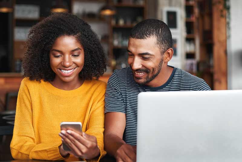 A man and woman sitting down with a computer Infront of him and both looking at a phone.