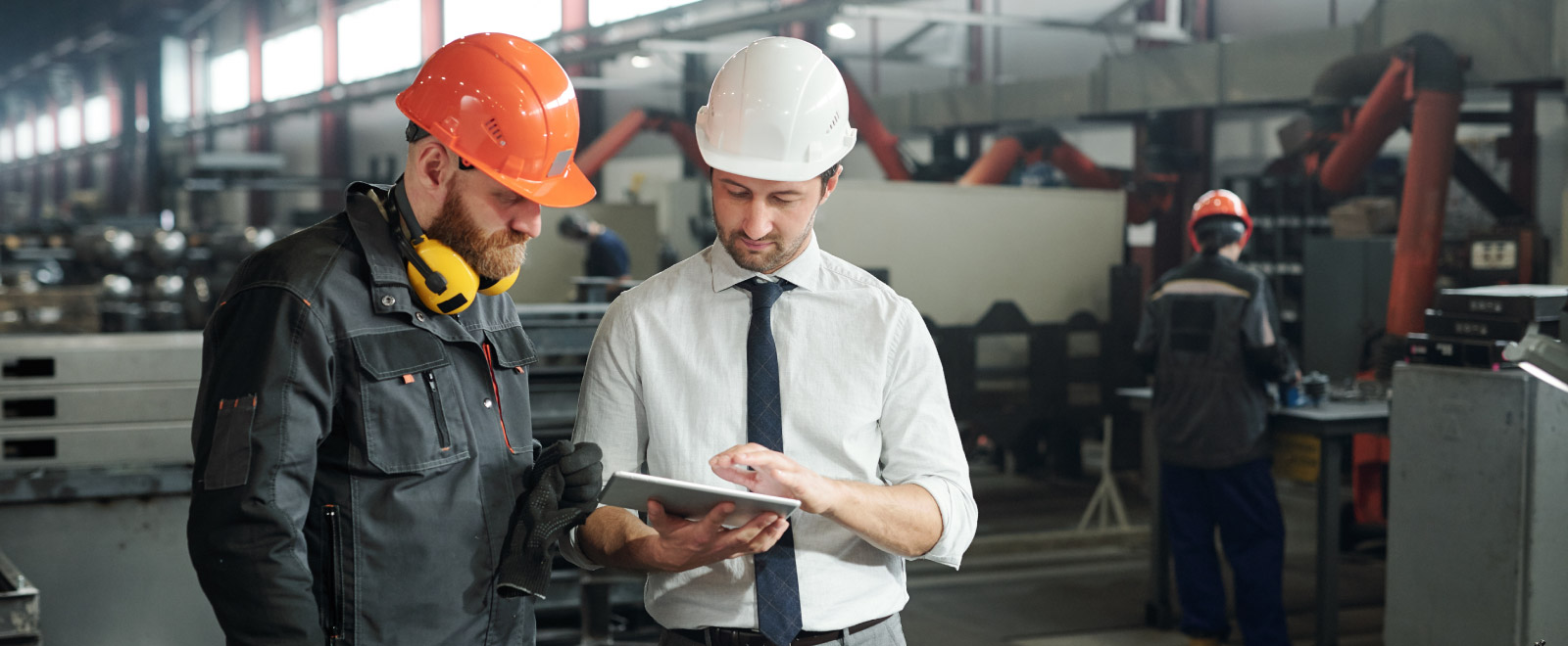 Two men wearing a hard hat looking at a tablet in a factory.