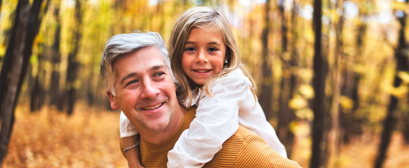 A little girl on her Dad's back in the woods smiling.
