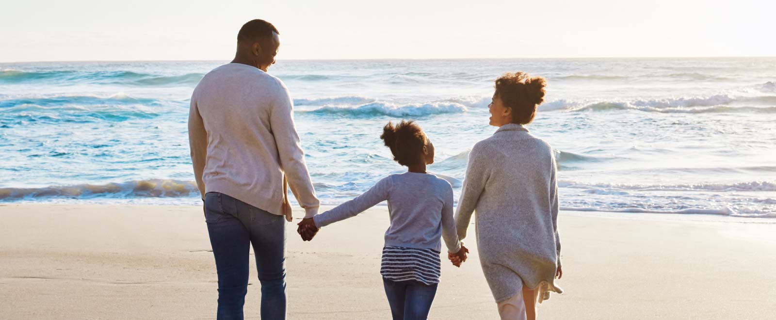 A family of three holding hands and walking towards the ocean.