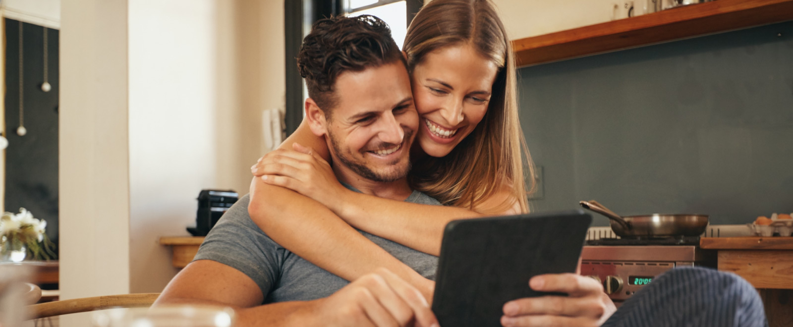 A man holding a tablet smiling with a woman hugging him from behind and looking at the tablet.