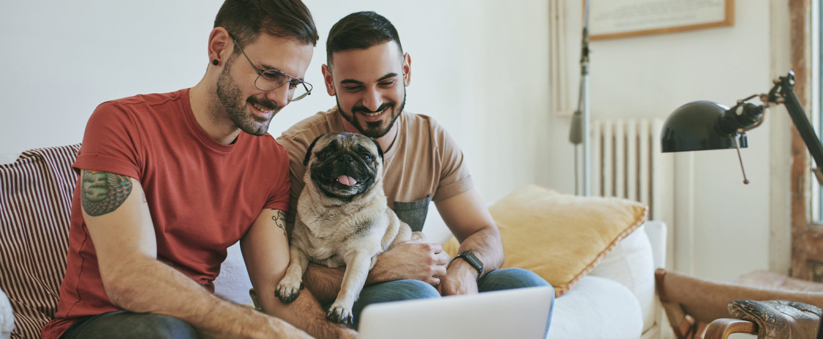 Two men sitting on a couch looking at a laptop with a pug.