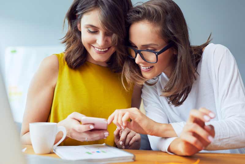 Two women sitting at a table looking at one of their cellphones.