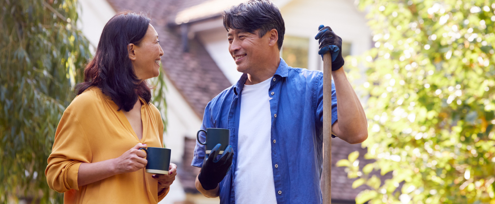 A couple smiling at one another, each holding a coffee mug and the man has on gloves and is holding up a shovel.