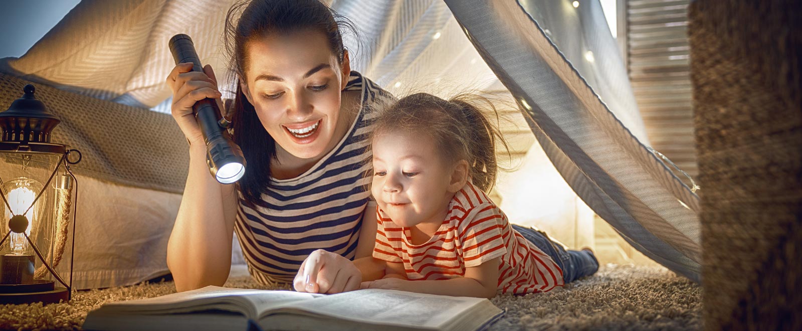 A woman and her daughter under a fort, reading with a flashlight.
