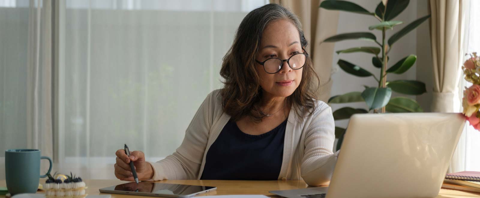 A woman wearing glasses looking at a laptop and hovering a stylus over a tablet.